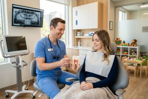 Smiling dentist explaining root canal therapy to a calm patient in a bright London, Ontario dental clinic with digital x-ray monitor and children's play area visible